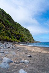 A rocky black sand beach beneath steep cliffs and blue sky