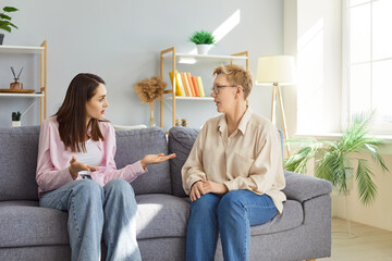 Young woman and senior woman sitting together on sofa at home, having serious conversation. Elderly mother listening to adult daughter expressing concern, sharing thoughts and actively gesticulating.