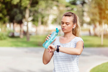 Young blonde woman jogging in park checking smartwatch and hydrating with water