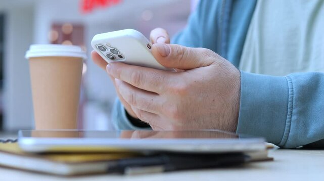 Man is using his cell phone while sitting at a table with a cup of coffee. The scene suggests a casual and relaxed atmosphere, as the man is focused on his phone rather than his surroundings