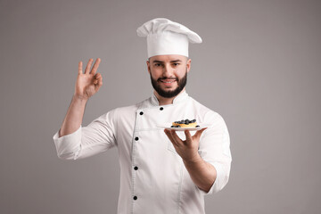 Happy confectioner in uniform holding delicious tart with blueberries and showing ok gesture on light grey background