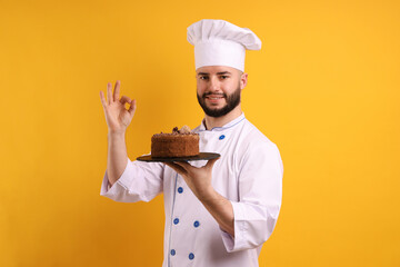 Happy confectioner in uniform holding chocolate cake and showing ok gesture on orange background