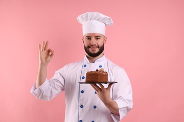 Happy confectioner in uniform holding chocolate cake and showing ok gesture on pink background