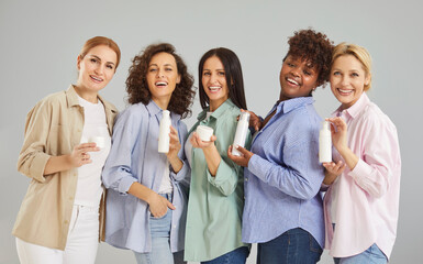 Portrait of happy smiling diverse women group in casual clothes looking cheerful at camera holding different facial and body cosmetics jars of cream. Beauty products and skin care concept.