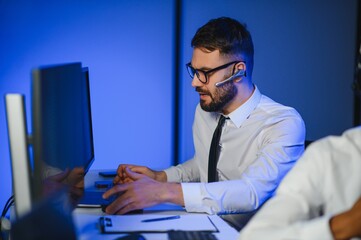 Happy Confident Technical Customer Support Specialist Having a Headset Call in a Monitoring and Control Room Filled with Colleagues and Display Screens