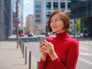 Fototapeta premium Beautiful woman in walking in Frankfurt on spring day and holding mobile phone. Urban lifestyle concept. Check social networks, send sms or book hotel, exuding confidence and professionalism