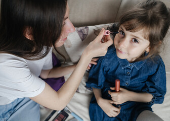 Mother putting makeup on her daughter face and they fool around together. Communication between women in the family. Fun time. Happy family concept. little girl doing her makeup