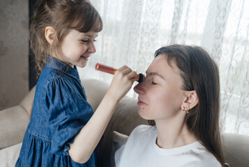 Daughter puts makeup on her mother face and plays around. Caring parent allows her child to paint her face. Happy childhood. Concept of loving family and fun. little girl doing her makeup