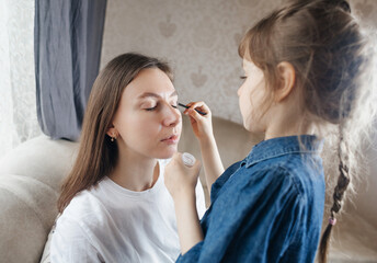 Daughter puts makeup on her mother face and plays around. Caring parent allows her child to paint her face. Happy childhood. Concept of loving family and fun. little girl doing her makeup