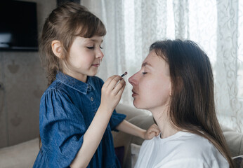 Daughter puts makeup on her mother face and plays around. Caring parent allows her child to paint her face. Happy childhood. Concept of loving family and fun. little girl doing her makeup