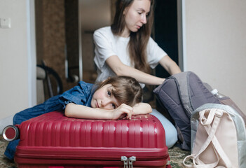 Tired 6 year girl lying on travel suitcase while mother is getting ready for trip. Cute child lost strength after packing bag and preparing for trip. Loving family, child care, home schooling concept