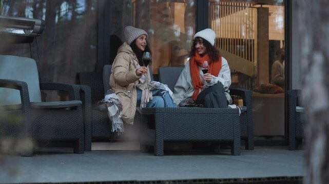 A European woman in her late 20s and a Latina woman in her 30s sitting on a sofa at a table with glasses of wine on a porch in front of a house with big windows and talking. dressed in beanies and jac