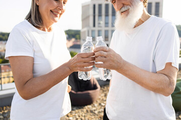 Joyful senior man and woman enjoying healthy lifestyle, drinking bottled water together outdoors. Depicts hydration, bonding, promoting wellness. Man has beard, both wear white shirts.