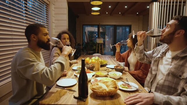 A group of four adult friends in their 30s, a Black couple, and a Caucasian couple, making a toast together with their wine glasses and chatting, as they're having dinner in a cozy house in the woods