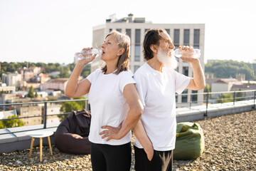 Older man and woman standing together outdoors, enjoying refreshing bottled water post workout. Rooftop setting with urban scenery creates healthy, active lifestyle atmosphere.