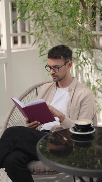 Man reading a book and drinking coffee in an outdoor cafe. He is wearing a beige shirt and glasses, sitting in a comfy chair.