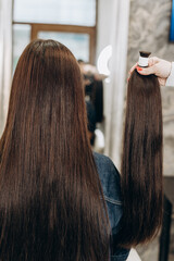 Fototapeta premium Hair stylist matching extensions to a woman’s long dark hair in a modern salon. Back view of hair lengthening procedure. Beauty treatment and professional haircare concept.