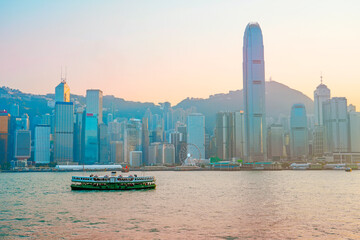 Fototapeta premium Classic green ferry at the pier on the route sails evening sunset view of tall glass skyscrapers in the financial district building, a symbol of the region's economic development. China, Hong Kong