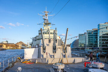 Turrets of HMS Belfast rise against the skyline of London. Visitors explore the ship while enjoying views of the River Thames and surrounding architecture on a clear day.