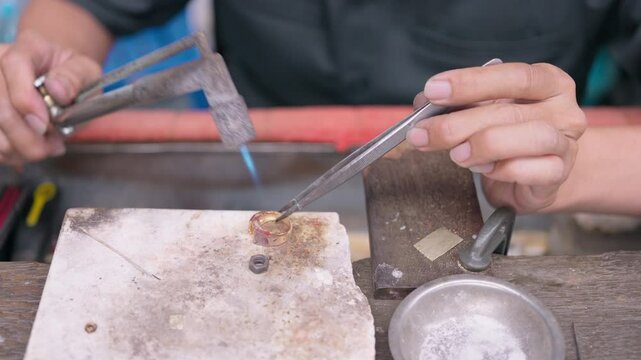 Close-up of a jeweler&rsquo;s hands using a blowtorch and tweezers to carefully solder a gold ring on a workbench. This image captures the intricate process of jewelry making, Bangkok, Thailand