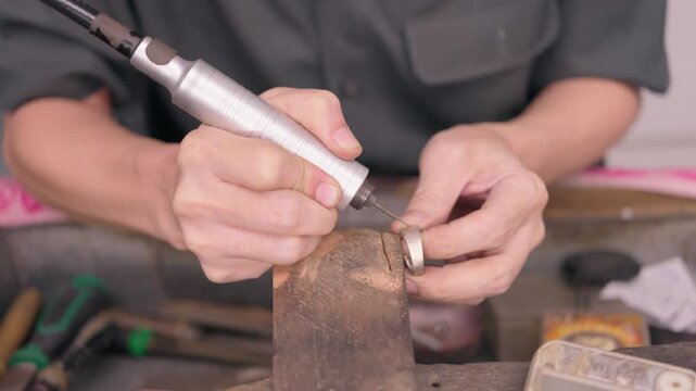 Close-up of a jeweller's hands engraving a gold and silver ring on a wooden block using a rotary tool. This image highlights the precision, craftsmanship and skill involved in customising, Bangkok 