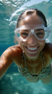 Portrait underwater of a smiling young woman wearing swim goggles and a bikini, looking directly at the camera while swimming in clear blue water. Generative AI