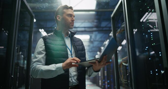 Male Data Center IT Engineer Standing in a Room with Server Racks. Cloud Computing Architect Using Laptop Computer for Servicing the System in Cyber Security and Data Protection Facility