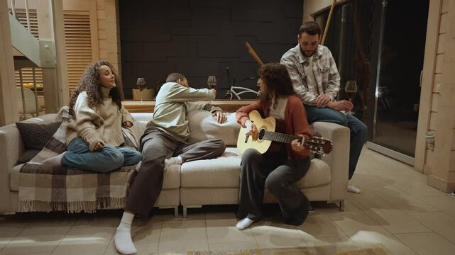 A multiethnic group of two men and one woman in their 30s sitting with glasses of wine on a white sofa around a White adult woman playing guitar and singing for them in a cozy living room