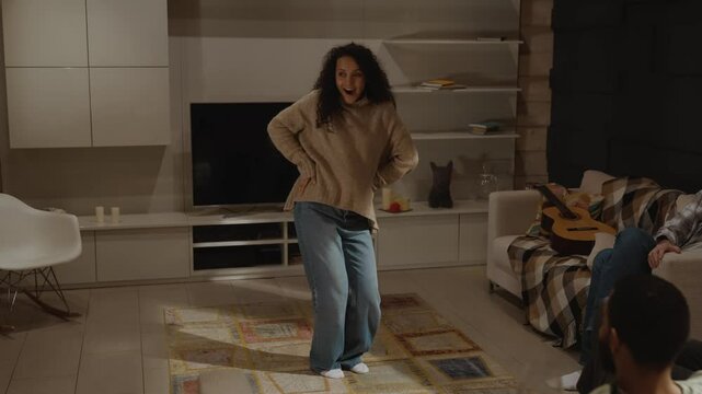 In the living room, a happy dark-haired Black woman in her early 30s is dancing the macarena and calling a European adult couple and her adult man to join her in enjoying.