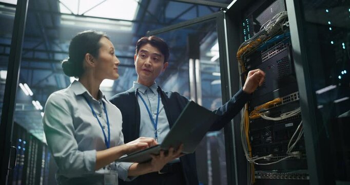 Portrait of Two Asian IT Engineers Standing in a Data Center Facility. Female and Male Technicians Using Laptop Computer while Discussing Work Beside an Open Server Rack Cabinet