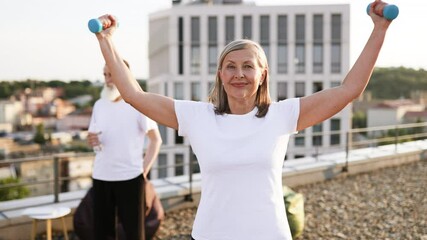 Older woman with gray hair wearing white shirt lifting dumbbell, outdoors, urban background. Happy and active lifestyle of senior adults, focusing on health, fitness, and positivity. - Powered by Adobe