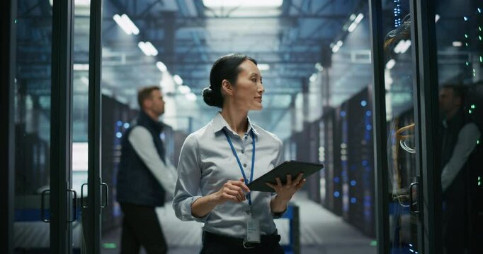 Portrait of an Asian Female IT Manager Using Tablet Computer, Walking Between Server Rack Cabinets and Supervising Data Center Operation. Male Pushing a Cart in the Background. Slow Motion Footage