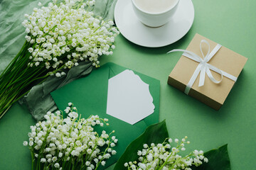 Morning coffee, gift and lily of the valley flowers with envelope on green background