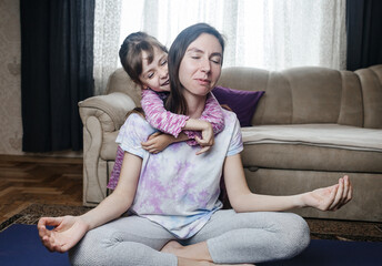 Young mother tries to meditate and do a home workout while her playful daughter gets in her way. Interrupted yoga at home and parenting reality. Mom and daughter having fun