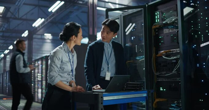 Asian Male Network Engineer and Female Data Analyst Collaborate on a Laptop Computer in a Modern Data Center Facility, Talking About Network Performance Optimization and Data Flow Across Servers