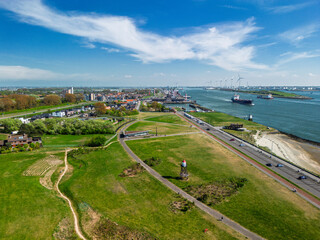 Aerial view of a coastal town with green parks, residential areas, a riverfront road, and busy shipping lanes. Wind turbines and industrial port facilities are visible across the water.