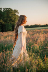 Brunette woman with long hair in a beige dress standing in a field at sunset, close-up portrait...