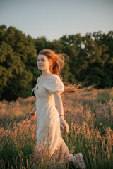 Brunette woman with long hair in a beige dress standing in a field at sunset, close-up portrait...