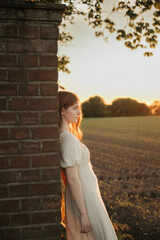 Brunette woman with long hair in a beige dress standing in a field at sunset, close-up portrait...