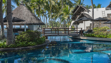 Obraz premium Swimming pool on a tropical resort. Clear water, blue tiled bottom, wooden pedestrian bridge. Ornamental plants in flower beds. Hotel buildings with thatched roofs, sun umbrellas, sun beds. Palm trees