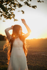 Brunette woman with long hair in a beige dress standing in a field at sunset, close-up portrait...