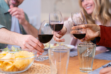 Close-up of unrecognizable happy friends toasting with red wine at a cheerful dinner party. Celebration, friendship, and good food in a warm and vibrant atmosphere at the table. 