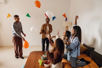A multiracial group of adult friends are playing with balloons during a party at home. Some sit on the couch, and others stand, enjoying the celebration indoors with snacks.