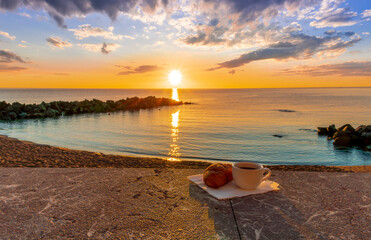 cup of coffee or tea with croissant on a morning embarkment beach with blue sea and beautiful cloudy sunrise or sunset on background, street food breakfast concept