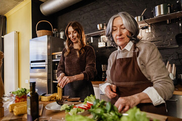 An elderly White woman and an adult woman prepare a meal together in a kitchen, cutting fresh vegetables and corn on a wooden table during a casual indoor cooking session.