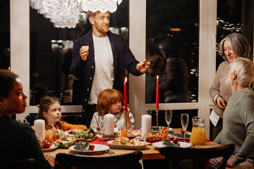 A multinational group of people of different ages is gathered around a dinner table at night, celebrating Christmas with candlelight, food, and conversation in a dining room with large windows.