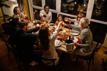 A diverse family of elderly individuals, adults, and children are clinking their glasses while sitting at a festive dinner table and smiling. A concept of celebration, reunion, and cuisine.