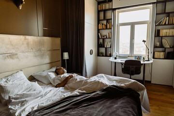 A senior White man peacefully sleeping on his back in a spacious bedroom with a large window, modern furniture, white bedding, and built-in black shelving filled with books and minimal decorations.