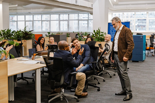 The multiracial adult men and women interact and applaud during a business meeting in a modern open-plan office with desks, laptops, and windows in the background.