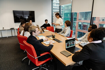 The multiracial adult men and women sit around a large conference table during a business meeting in a modern office, with one woman standing and presenting to the group.